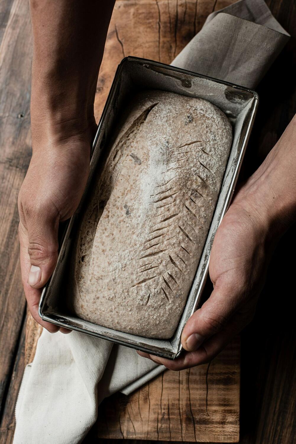 Two hands hold a metal loaf pan with a scored, unbaked bread loaf above a wooden table
